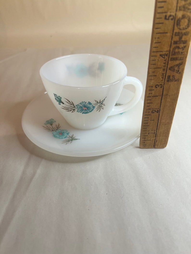 May include: A white teacup and saucer set with a floral design featuring blue flowers and green leaves.
