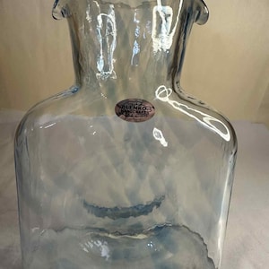 May include: A clear, light blue glass pitcher with a square base and a wide, flared top. The pitcher has a textured surface and a small, round sticker that reads "Bluenko Handcrafts".