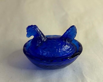 May include: A cobalt blue hen-shaped trinket dish. The dish features a detailed hen figure atop a textured bowl. The glass has a glossy finish, and the design is reminiscent of vintage glassware.