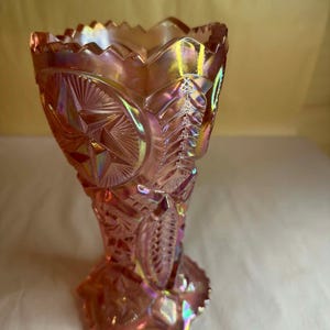 May include: A pink iridescent glass vase with intricate embossed patterns. The vase features a starburst design, leaf motifs, and a ruffled rim. The glass has a shimmering, opalescent quality, reflecting various colors.