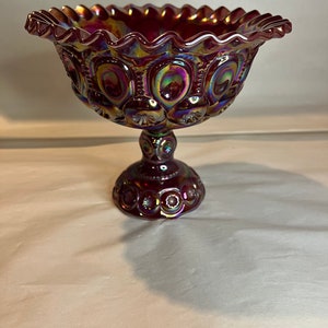 May include: A ruby red, iridescent glass pedestal bowl. The bowl has a ruffled edge and is decorated with a pattern of circles and crescent shapes. The bowl is approximately 7 inches in diameter and 6 inches tall.