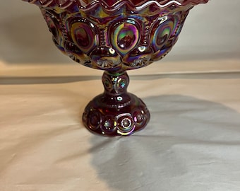 May include: A ruby red, iridescent glass pedestal bowl. The bowl has a ruffled edge and is decorated with a pattern of circles and crescent shapes. The bowl is approximately 18 cm in diameter and 15 cm tall.