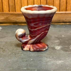 May include: A decorative glass cornucopia vase with a red, white, and purple swirled pattern. The vase has a flared opening with a scalloped rim and a curled horn-like design. The base is a rounded shape.