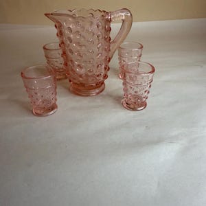 May include: A set of five pink glass cups and a jug with a textured bubble design. The jug has a handle and a scalloped rim.