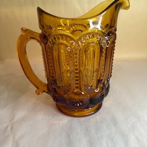 May include: Amber-colored glass pitcher with a decorative pattern. The pitcher has a handle and a spout for pouring. The glass is a warm, golden color, and the design includes vertical and arched elements. The base has a starburst design.