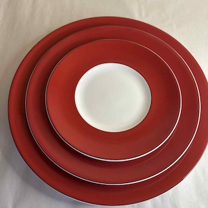Puede incluir: Una pila de tres platos de cerámica rojos con un centro blanco. Los platos están anidados, con una fina línea blanca que separa las secciones rojas y blancas. Los platos son de color rojo liso.