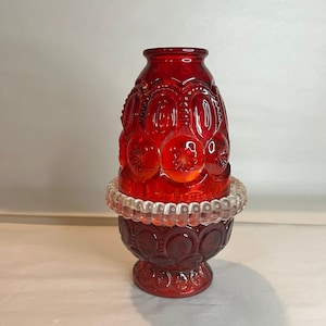 Pode incluir: Um castiçal de vidro vermelho rubi com um design de estrelas e luas. O castiçal tem uma base redonda, uma faixa de contas de vidro transparente e um topo alargado. O vidro é de cor vermelha profunda e o design é em relevo.