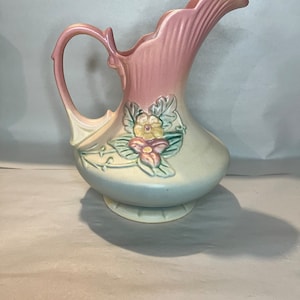 Puede incluir: Una jarra de cerámica vintage con un esmalte rosa y azul. La jarra presenta un diseño floral con flores rosas, amarillas y verdes.