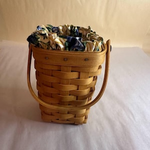 May include: A woven basket with a wooden handle. The basket is rectangular with a rounded top and is lined with a floral fabric. The fabric features a pattern of yellow, blue, and green flowers. The handle is made of a curved piece of wood.
