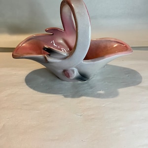 Puede incluir: Una cesta de cerámica blanca y rosa con asa. La cesta tiene forma de cisne con cuello largo y asa curva.