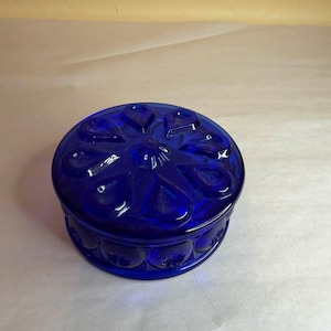 Könnte beinhalten: Eine runde, kobaltblaue Glas-Schmuckschatulle mit Deckel. Der Deckel hat ein Blumenmuster, und die Seiten der Schatulle sind mit einem sich wiederholenden Muster aus kleinen Kreisen verziert. Die Schatulle steht auf einer weißen Oberfläche.