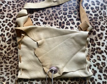 May include: A light beige leather crossbody bag with a flap closure and a decorative metal button. The bag has a long strap and a braided detail on the side.