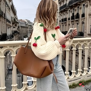 May include: Cream-colored knit sweater adorned with red cherry appliqués and green leaves. A large brown tote bag hangs from the shoulder. The person is wearing light blue jeans and is holding a phone. The background shows a European city street.