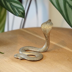 May include: A brass cobra ornament with a detailed, textured finish. The snake is coiled on a wooden surface, with its head raised. A decorative item.