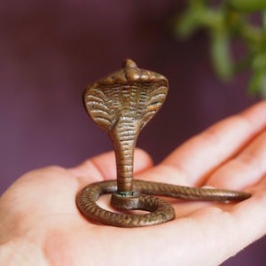 May include: A small, bronze-coloured cobra statue with its head raised and its body curled in a circle. The statue is resting on a person's hand.