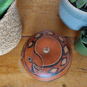 Può includere: Una zucca marrone fatta a mano con un disegno di serpente intagliato e dipinto di nero. La zucca è circondata da piante in vaso e un cesto intrecciato su una superficie di legno. Il disegno del serpente è il punto focale.