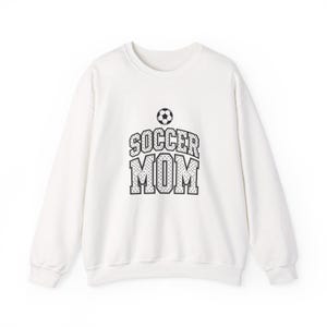 May include: A white long-sleeve crewneck sweatshirt featuring a black graphic print on the chest. The design includes a small soccer ball icon above the text SOCCER MOM, written in a bold, block-style font with a dotted pattern inside the letters.