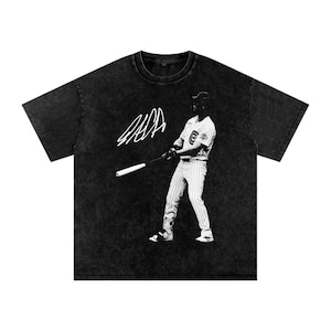 Może przedstawiać: Czarny t-shirt z efektem sprania kwasem, ozdobiony białym nadrukiem graficznym przedstawiającym baseballistę w prążkowanym stroju zamachującego się kijem. Nad postacią znajduje się biały, stylizowany podpis. Koszulka ma okrągły dekolt i krótkie rękawy.