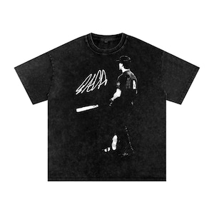 Puede incluir: Una camiseta de manga corta negra con lavado ácido que presenta un estampado gráfico blanco de un jugador de béisbol con gorra sosteniendo un bate. A la izquierda del jugador hay una firma blanca grande y estilizada. Cuello redondo.