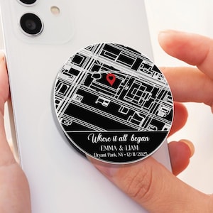 以下が含まれることがあります： 白のスマートフォンと白黒の地図デザインのスマホグリップ。 グリップには赤いハートと「Where it all began」の文字、名前と日付が記されています。 電話は人の手に持たれています。