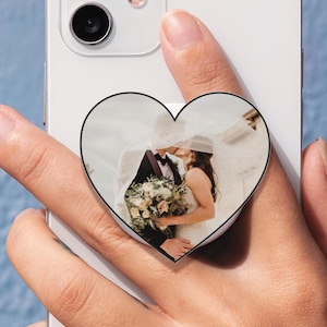 May include: A white smartphone with a heart-shaped phone grip featuring a wedding photo. The grip shows a couple kissing, with the bride holding a bouquet. The phone is held against a light blue background.