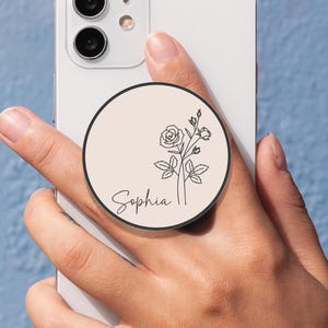May include: A white smartphone with a black camera module is held in a hand. A round phone grip is attached to the phone. The grip has a light beige background with a black outline. A black floral design and the name "Sophia" are printed on the grip.