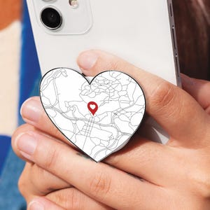 May include: A white smartphone with a heart-shaped phone grip featuring a black and white map design and a red location marker. The phone is held in a person's hand.