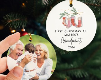 May include: Two round Christmas ornaments. One features a family photo, the other has a festive design with stockings and text that reads "First Christmas as Matteo's Grandparents 2026". Both ornaments are white with gold hanging cords.