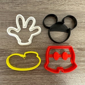 May include: Four plastic cookie cutters in the shape of Mickey Mouse's head, hand, shorts, and a single shoe. The cutters are white, black, red, and yellow.
