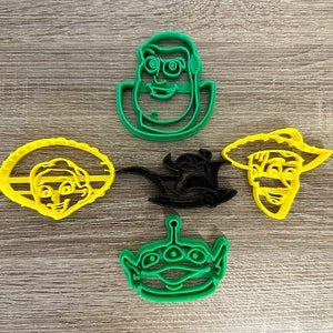 May include: Five plastic cookie cutters shaped like characters from the Toy Story movie franchise. The cutters are green, yellow, and black. The characters include Woody, Buzz Lightyear, Jessie, Rex, and Alien.