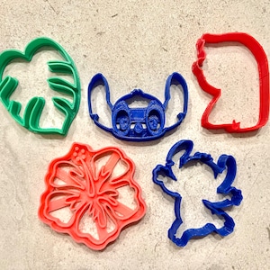 May include: Five cookie cutters in various shapes and colors. The cutters are shaped like a monstera leaf, a hibiscus flower, and three different Stitch characters from the Disney movie Lilo & Stitch.