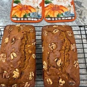 May include: Two loaves of freshly baked walnut bread sit on a black wire rack, with two pumpkin-themed pot holders in the background. The pot holders have an orange border and the word "thankful".
