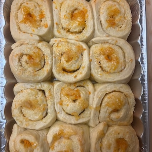 May include: A baking pan filled with orange rolls, each roll is topped with a sweet orange glaze and orange zest.