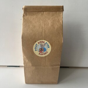 May include: A brown paper bag with a yellow circle sticker featuring a cartoon baker with a red beard and blue shirt. The sticker says "Polly Anna Star Bakery".