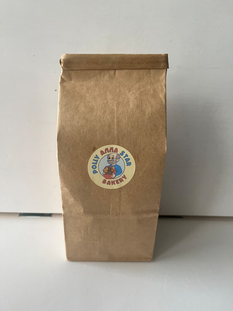 May include: A brown paper bag with a yellow circle sticker featuring a cartoon baker with a red beard and blue shirt. The sticker says "Polly Anna Star Bakery".