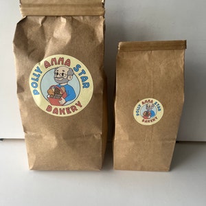 May include: Two brown paper bags with a logo for Polly Anna Star Bakery. The logo features a cartoon man with glasses holding a basket of bread. The text "Polly Anna Star Bakery" is in a circle around the cartoon.