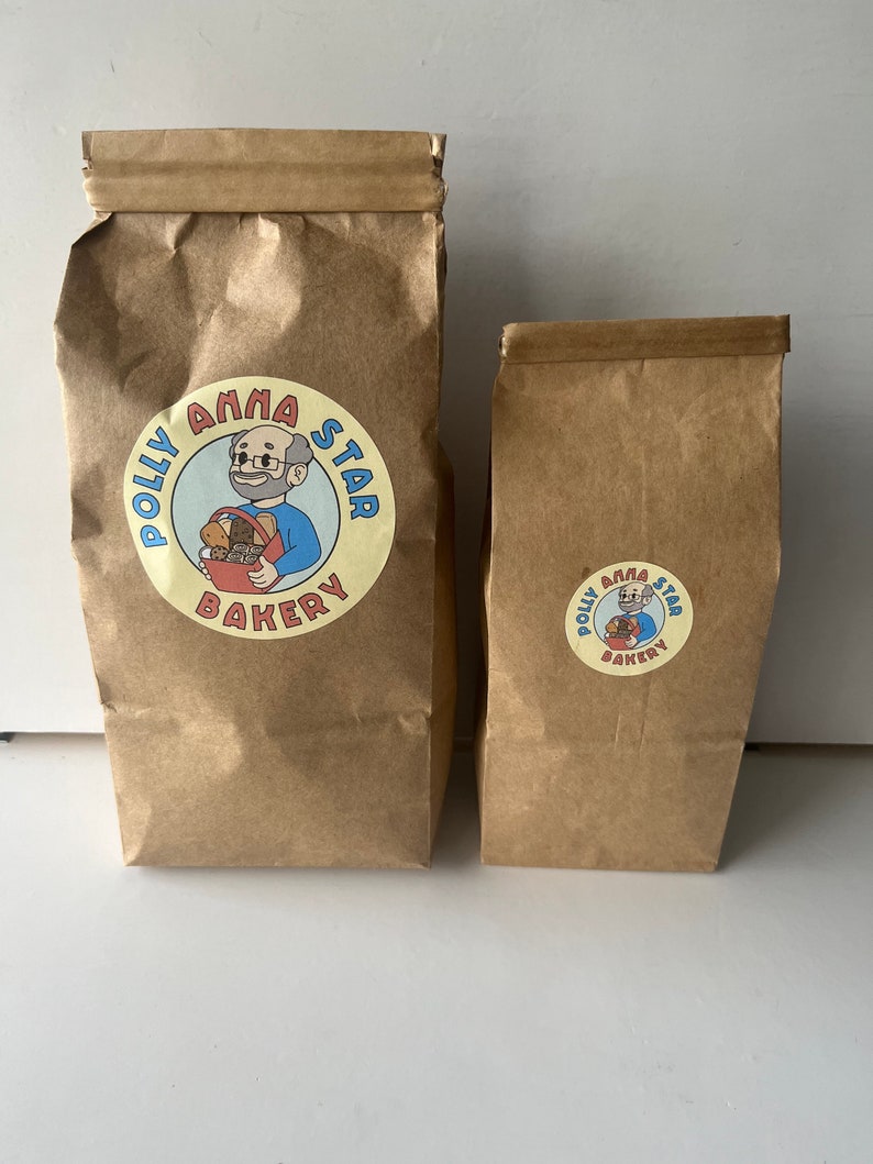May include: Two brown paper bags with a logo for Polly Anna Star Bakery. The logo features a cartoon man with glasses holding a basket of bread. The text "Polly Anna Star Bakery" is in a circle around the cartoon.