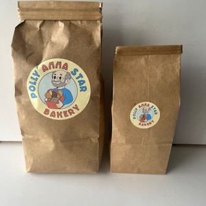 May include: Two brown paper bags with a logo for Polly Anna Star Bakery. The logo features a cartoon man with glasses and a red shirt holding a basket of bread. The text "Polly Anna Star Bakery" is in a circle around the cartoon.