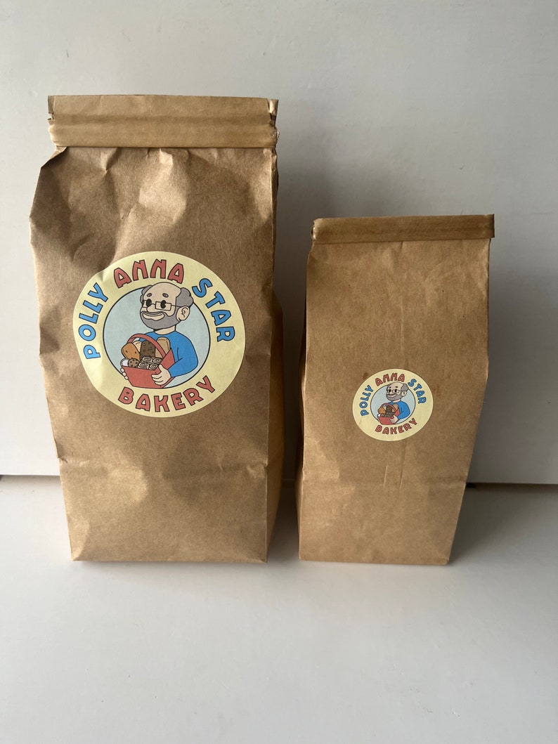 May include: Two brown paper bags with a logo for Polly Anna Star Bakery. The logo features a cartoon man with glasses and a red shirt holding a basket of bread. The text "Polly Anna Star Bakery" is in a circle around the cartoon.