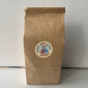 May include: A brown paper bag with a round sticker featuring a cartoon illustration of a person holding a basket of baked goods. The sticker says "Polly Anna Star Bakery".
