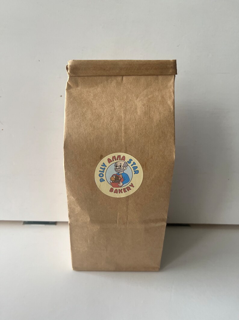 May include: A brown paper bag with a round sticker featuring a cartoon illustration of a person holding a basket of baked goods. The sticker says "Polly Anna Star Bakery".