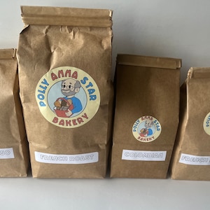 May include: Four brown paper bags with white labels. The bags are labeled with different coffee blends: Espresso, French Roast, Colombian, and French Roast. Each bag has a logo with a cartoon baker and the text "Polly Anna Star Bakery".