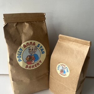 May include: Two brown paper bags with the Polly Anna Star Bakery logo. The logo features a cartoon man holding a basket of bread. The larger bag is standing upright, while the smaller bag is lying on its side.