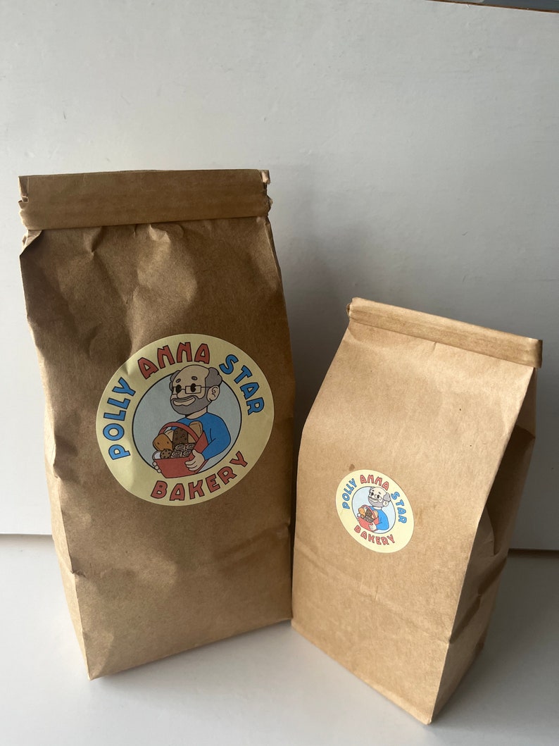 May include: Two brown paper bags with the Polly Anna Star Bakery logo. The logo features a cartoon man holding a basket of bread. The larger bag is standing upright, while the smaller bag is lying on its side.