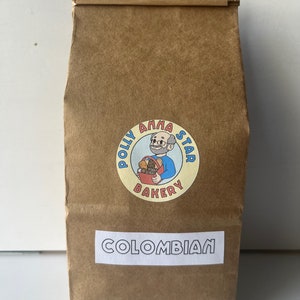 May include: A brown paper bag with a sticker featuring a cartoon character holding a basket of baked goods. The sticker says "Polly Anna Star Bakery". The bag also has a white label that says "COLOMBIAN".