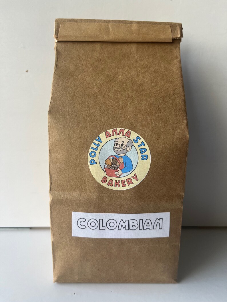 May include: A brown paper bag with a sticker featuring a cartoon character holding a basket of baked goods. The sticker says "Polly Anna Star Bakery". The bag also has a white label that says "COLOMBIAN".