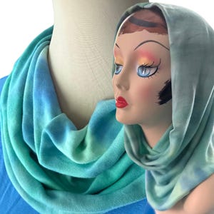 May include: A soft, tubular infinity scarf featuring a tie-dye pattern in shades of teal, turquoise, and light blue. The fabric is draped in loose folds around a mannequin head and neck, displaying a smooth, consistent texture throughout the piece.