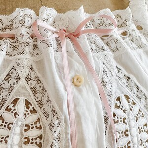 May include: Close-up of a white vintage blouse with intricate lace detailing. The blouse features a delicate pink ribbon tied in a bow at the neckline and a single button closure. The lace has a floral pattern.