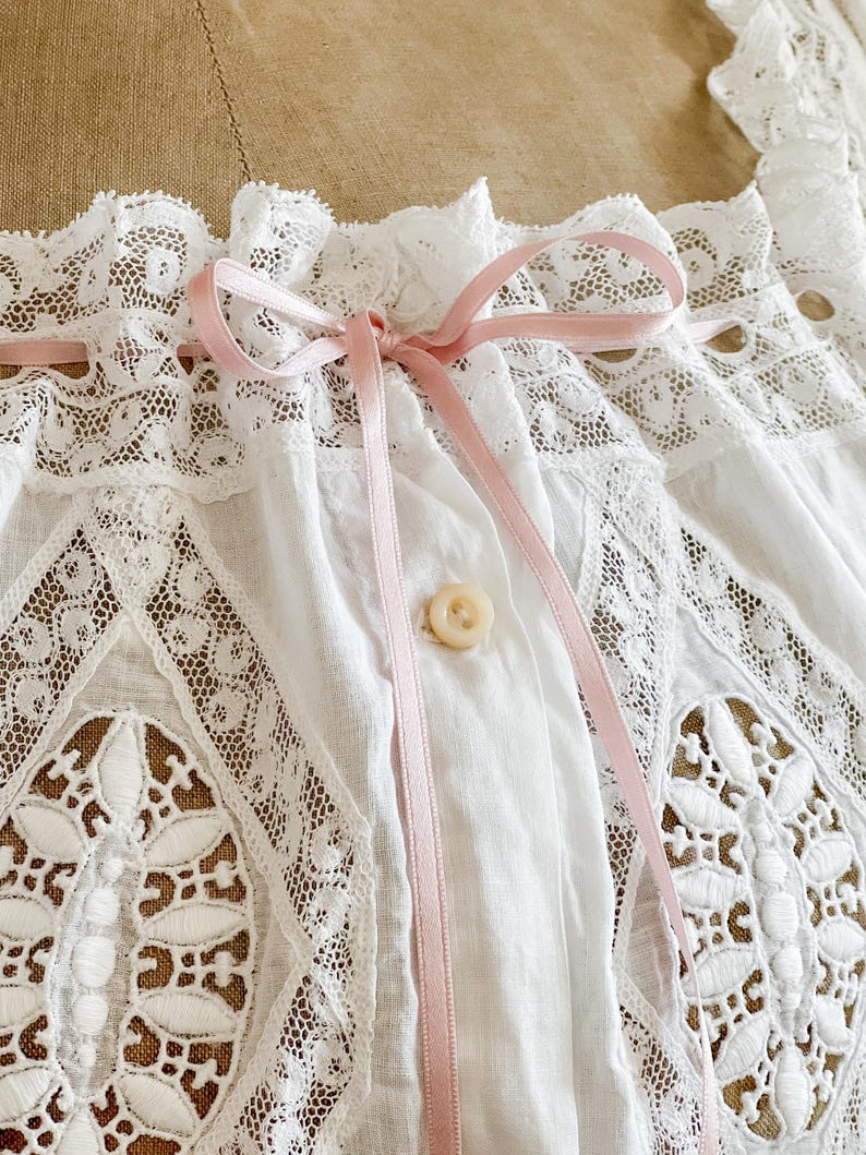 May include: Close-up of a white vintage blouse with intricate lace detailing. The blouse features a delicate pink ribbon tied in a bow at the neckline and a single button closure. The lace has a floral pattern.