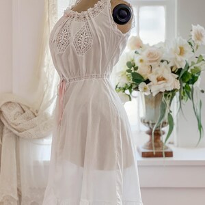 May include: A white vintage-style nightgown on a mannequin. The sleeveless gown features lace detailing on the bodice and hem, with a pink ribbon accent at the waist. The dress is displayed in front of a window with a floral arrangement.